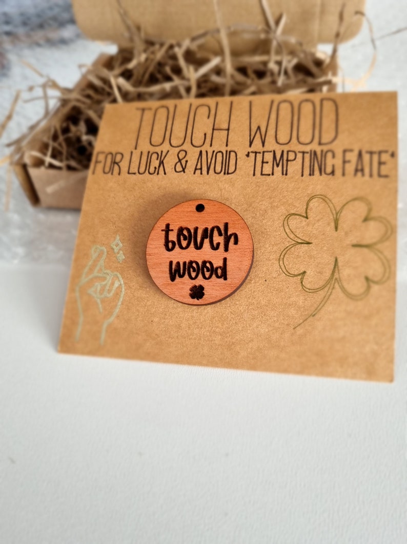 May include: A round wooden charm with the text "touch wood" and a four-leaf clover symbol. The charm is on a brown card with the text "Touch Wood for Luck & Avoid 'Tempting Fate'".