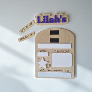 May include: A wooden milestone board with the name "Lilah's" in purple. Includes labels for "Last Day Of" and "First Day Of". The board has sections for date, things I love, and "When I grow up I want to be".