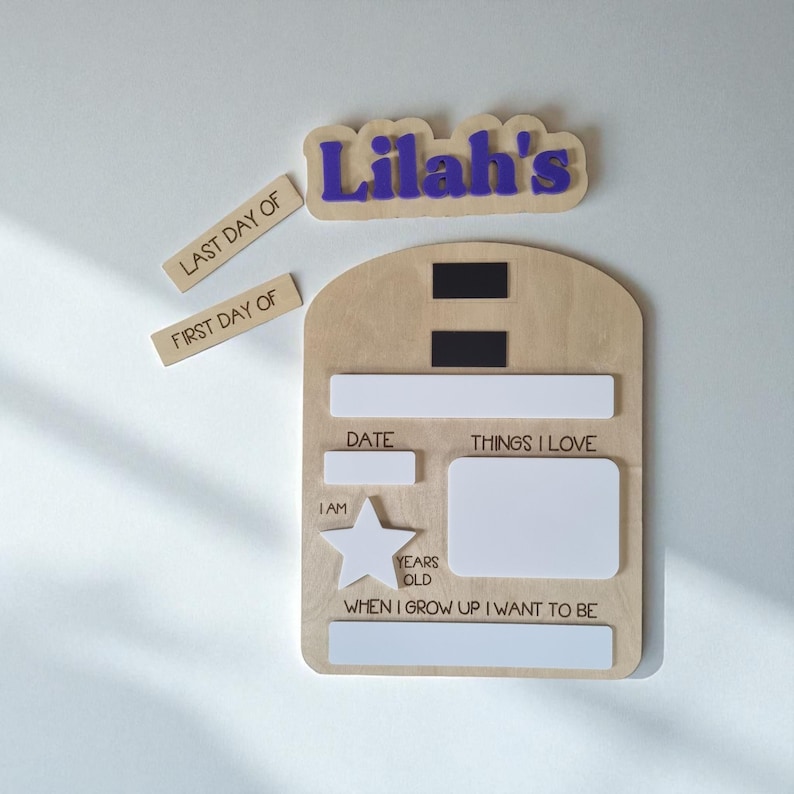 May include: A wooden milestone board with the name "Lilah's" in purple. Includes labels for "Last Day Of" and "First Day Of". The board has sections for date, things I love, and "When I grow up I want to be".