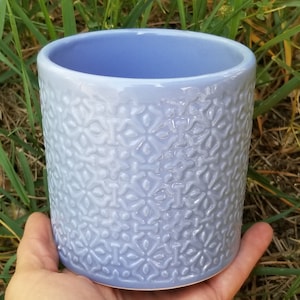 May include: A light blue ceramic planter with a white and light blue embossed pattern. The planter has a cylindrical shape and a smooth, glazed finish.