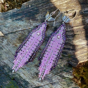 Op de afbeelding: Een paar lange, roze en paarse kralen oorbellen met vlinder bedels. De oorbellen zijn gemaakt van kleine, kleurrijke kralen en hebben een delicate, vrouwelijke uitstraling.