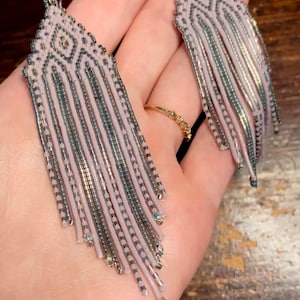 May include: A pair of long, pink and silver beaded earrings with a geometric pattern. The earrings have a fringe design and are hanging from a hook.