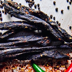 May include: Close-up of a pile of dark, dried meat strips with a spicy, peppery flavor. The meat is covered in black peppercorns and red pepper flakes.
