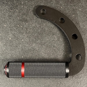 May include: Black metal handle with a curved design and three circular holes. The handle features a textured black grip with a red stripe and a rounded end with embossed text. The item is designed for fitness equipment.
