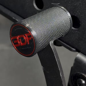 May include: A black cylindrical gym equipment attachment with a textured knurled surface and a flat circular end cap featuring the red text 3DF. The component is mounted to a black metal frame with circular cutouts and a large bolt head visible.