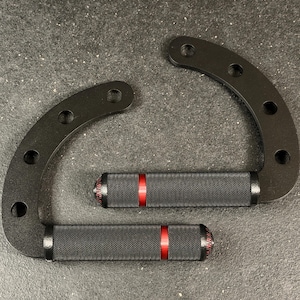 May include: Two black, curved metal handles with textured, black grips and a red stripe. Each handle has multiple holes for mounting. The handles are designed for a specific purpose, likely for exercise equipment or a similar application.