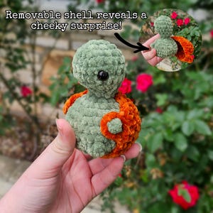 May include: A handmade, green and orange crochet creature with a black eye. The text "Removable shell reveals a cheeky surprise!" is above the creature. The creature is held in a hand against a background of green foliage and red flowers.