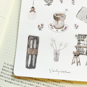 May include: A set of watercolor stickers featuring a variety of images, including a cup of coffee, a vase of flowers, a pen holder, a light fixture, and a table and chair. The stickers are in shades of brown, gray, and white.
