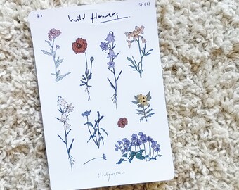 Pode incluir: Uma folha de adesivos com ilustrações desenhadas à mão de várias flores silvestres. Os adesivos são brancos com contornos pretos e apresentam flores em tons de vermelho, amarelo, azul e rosa. A folha é etiquetada como "Wild Flowers" e "SH003".