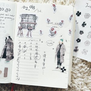 May include: A page from a planner with the date 02.08 written at the top. The page is divided into sections with handwritten notes and drawings. The notes include the words "to do", "but first coffee", and "Today". There are also several stickers on the page, including a small cafe scene, a person in a coat, and a few flowers.