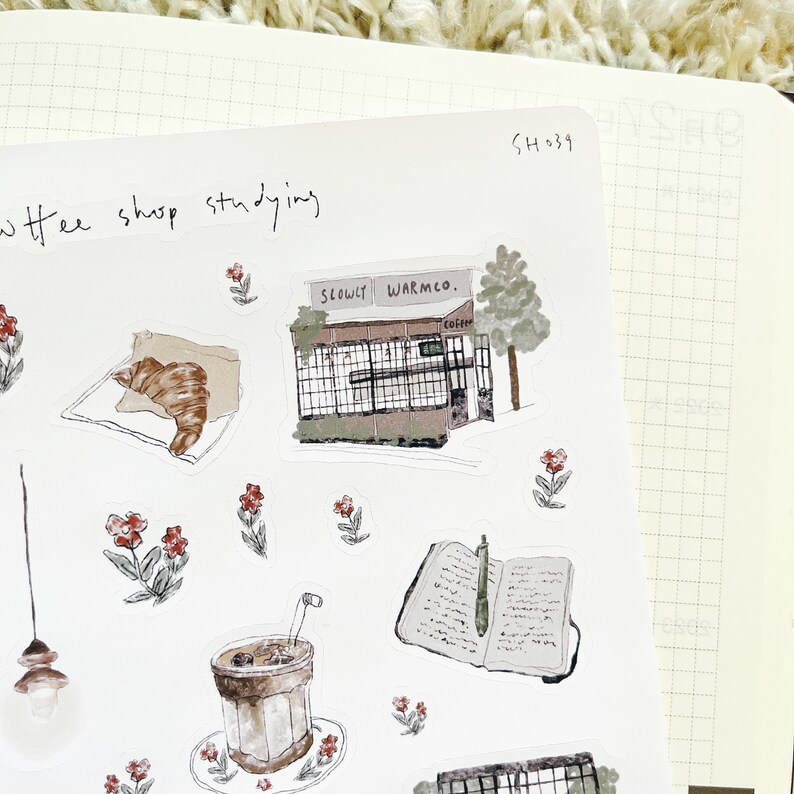 May include: A set of stickers featuring illustrations of a coffee shop, a croissant, a cup of coffee, a book, and flowers. The stickers are on a white background with a grid pattern.