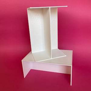 May include: A white, geometric display stand with a layered design. The stand has a rectangular base and an upright section with two vertical dividers. The background is a solid magenta color.