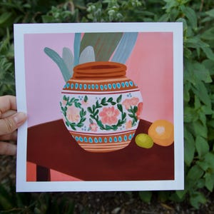 May include: A hand-painted ceramic vase with floral designs in shades of pink, green, and blue. The vase is sitting on a table with two small fruit, one orange and one green.