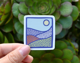 May include: A rectangular sticker with a landscape design. The sticker features a blue border, a light blue sky with a yellow sun, and rolling hills in shades of orange, green, and blue. The design has a hand-drawn aesthetic.