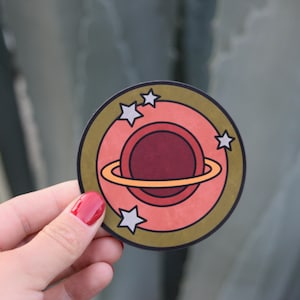 May include: A round sticker with a gold outline and a pink planet with a yellow ring. The planet has a red center and is surrounded by white stars.