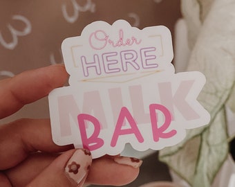 Puede incluir: Pegatina rosa y blanca con el texto "Order Here Milk Bar" en una fuente de estilo neón.