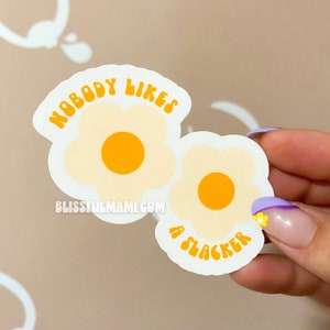 May include: Two white daisy stickers with yellow centers. The top sticker has the text "NOBODY LIKES" in orange. The bottom sticker has the text "A SLACKER" in orange.
