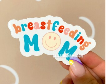 May include: A white sticker with the text "breastfeeding MOM" in colorful letters. A smiley face is in the middle of the word MOM. The sticker is being held by a hand with colorful nail polish.