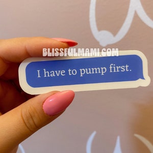 May include: A blue and white sticker with the text "I have to pump first." The sticker is held by a hand with pink nail polish. The website "BLISSFULMAMI.COM" is printed at the top of the sticker.