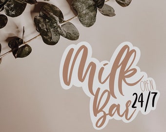 May include: A sticker with the words "Milk Bar" in a cursive font, with "Open 24/7" below it. The sticker is light brown with a white border, set against a beige background. A sprig of green leaves is in the upper left corner.
