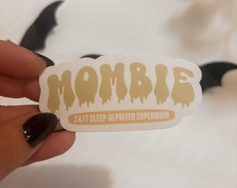 Puede incluir: Una pegatina beige con la palabra "MOMBIE" en una fuente que gotea, con el texto "24/7 SLEEP-DEPRIVED SUPERMOM" debajo. El texto es marrón. Decoraciones de murciélagos negros en el fondo.