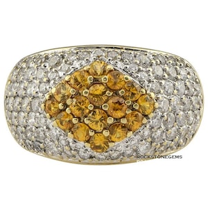 Może przedstawiać: Złoty pierścionek z dużym, diamentowym kamieniem centralnym otoczonym mniejszymi, okrągłymi kamieniami. Kamień centralny jest jasnożółty, podczas gdy otaczające go kamienie są przezroczyste. Pierścionek osadzony jest w obrączce ze złota.