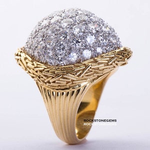 May include: A gold ring with a large, round, dome-shaped center set with many small diamonds. The band of the ring is textured and has a decorative design.