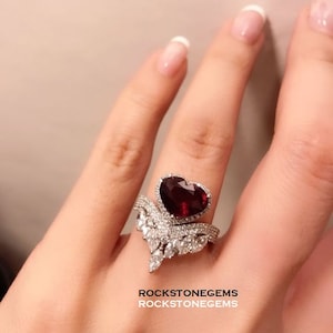 May include: A heart-shaped ruby ring with a white gold band and diamond accents. The ring is set in a delicate, V-shaped design. The ring is on a finger with a white manicure. The text "ROCKSTONEGEMS" is visible on the bottom of the image.