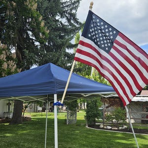 May include: An American flag with red and white stripes and a blue field of white stars is displayed outdoors. The flag is on a wooden pole, waving in the wind. A blue canopy and green grass are in the background.
