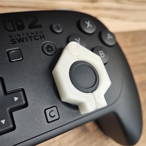 Peut inclure: Une manette Nintendo Switch noire avec un cache de joystick blanc et géométrique. La manette comporte divers boutons, dont un pavé directionnel, et le logo Nintendo Switch est visible. Le cache de joystick a un centre circulaire.