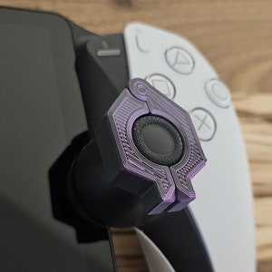 May include: A close-up of a purple and black gaming controller attachment. The attachment is a textured, hexagonal shape with a circular button in the center.
