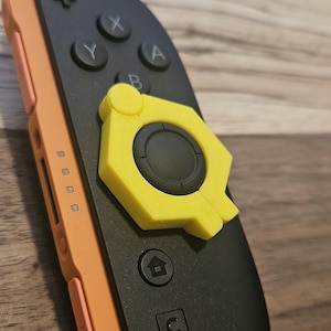May include: Black and orange video game controller with yellow accents. The controller has buttons labeled X, Y, A, and B, along with a home button and a C button. The controller is set against a wooden background.