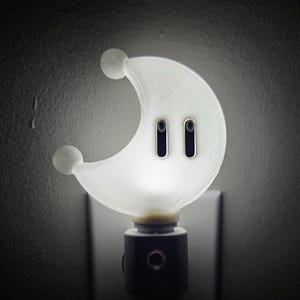 May include: A white crescent moon-shaped nightlight with two black eyes. The nightlight plugs into a standard electrical outlet.