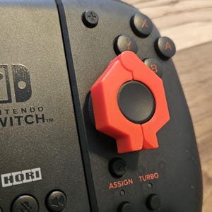 May include: A black Nintendo Switch controller with a red and black joystick attachment. The controller has the Nintendo Switch logo and the text "INTENDO SWITCH TM" on the front. The controller also has the text "HORI" on the front. The controller has buttons labeled "ASSIGN" and "TURBO".