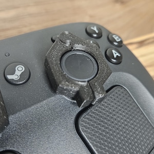 May include: Close-up of a black gaming controller with a custom 3D-printed thumbstick attachment. The controller has a Steam logo and A, B, and Y buttons. The thumbstick attachment is black and has a textured design.