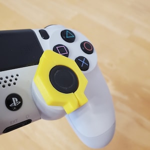 Könnte beinhalten: Nahaufnahme eines weißen und schwarzen PlayStation 4-Controllers mit einem gelben Zubehörteil. Das Zubehör hat eine sechseckige Form mit einem schwarzen Mittelteil. Die Tasten des Controllers sind blau, rosa, grün und rot.