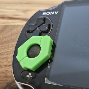 May include: A black Sony PlayStation Vita handheld gaming console with a green plastic accessory attached to the top right corner. The accessory has a circular opening in the center.