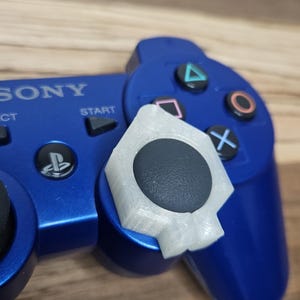 Op de afbeelding: Een blauwe Sony-gamecontroller met een donkergrijze analoge stick en een wit, halfdoorzichtig, zeshoekig plastic opzetstuk dat over de basis is geplaatst. De controller bevat standaardknoppen met driehoek-, cirkel-, kruis- en vierkantsymbolen.