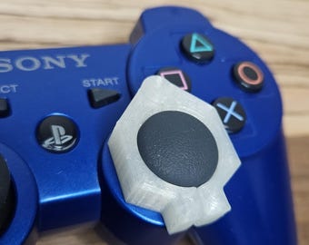 May include: A blue Sony game controller featuring a dark gray analog stick with a white, semi-translucent, hexagonal plastic attachment fitted over the base. The controller includes standard buttons labeled with triangle, circle, cross, and square symbols.