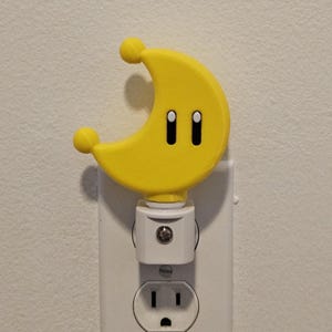 May include: A yellow crescent moon-shaped nightlight with two black eyes plugged into a white electrical outlet.