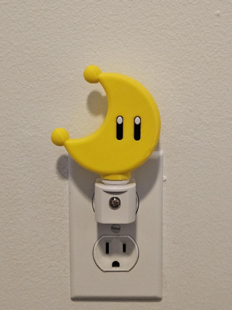 May include: A yellow crescent moon-shaped nightlight with two black eyes plugged into a white electrical outlet.