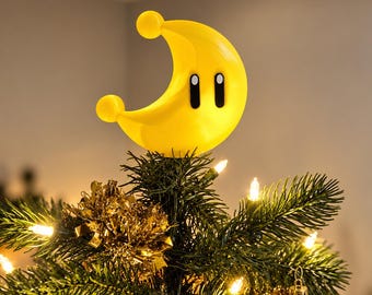 May include: A bright yellow crescent moon tree topper featuring two black oval eyes with white pupils. The moon has a smooth, rounded surface with a small sphere at each tip, positioned at the very top of a green pine Christmas tree.