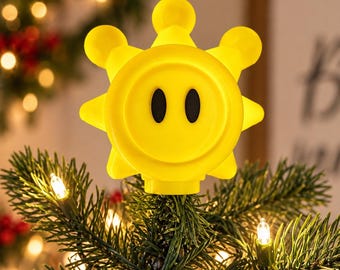 May include: A bright yellow sun-shaped tree topper featuring a stylized face with two black oval eyes. The object has a circular center with radiating rounded points and is positioned at the top of a green pine tree with small warm white lights.