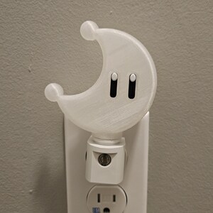 May include: A white crescent moon-shaped nightlight plugged into a standard electrical outlet. The nightlight has two small black buttons on the front.