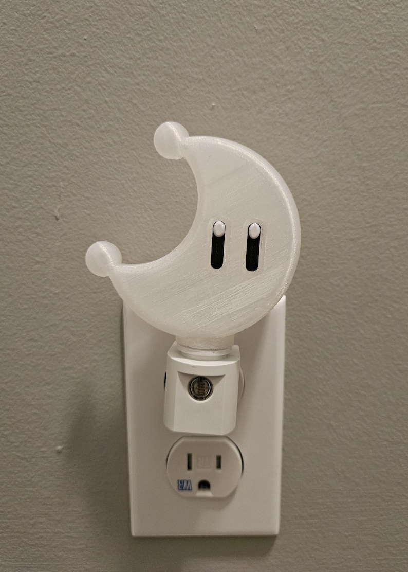 May include: A white crescent moon-shaped nightlight plugged into a standard electrical outlet. The nightlight has two small black buttons on the front.