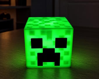 May include: A glowing green cube-shaped lamp featuring the pixelated face of a Creeper character. The light emits a bright neon green hue, highlighting the dark black square eyes and the inverted T-shaped mouth design on the front surface.