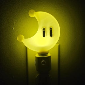May include: A yellow crescent moon shaped night light plugged into an electrical outlet. The night light has two small black circles on the front.