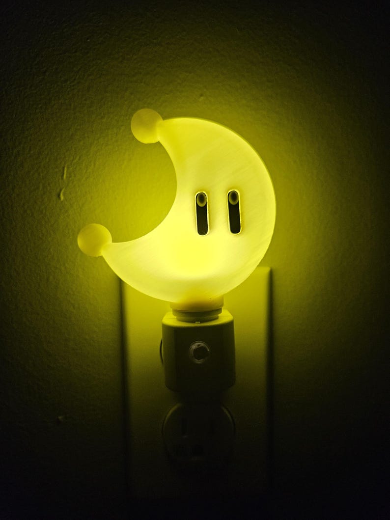 May include: A yellow crescent moon shaped night light plugged into an electrical outlet. The night light has two small black circles on the front.