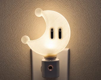 May include: A white, crescent moon-shaped night light plugged into a wall outlet. The moon features two small spheres at its tips and two vertical, black, oval-shaped eyes. The light emits a warm, soft glow, illuminating the textured wall surface.