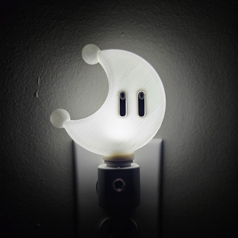 May include: A white crescent moon-shaped nightlight with two black eyes. The nightlight plugs into a standard electrical outlet.
